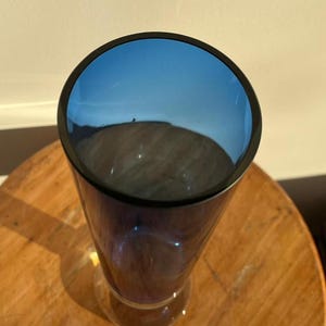 Op de afbeelding: Een hoge, heldere glazen vaas met een uitlopende voet en een donkerblauwe rand. De binnenkant van de vaas is diepblauw. De vaas staat op een houten oppervlak.