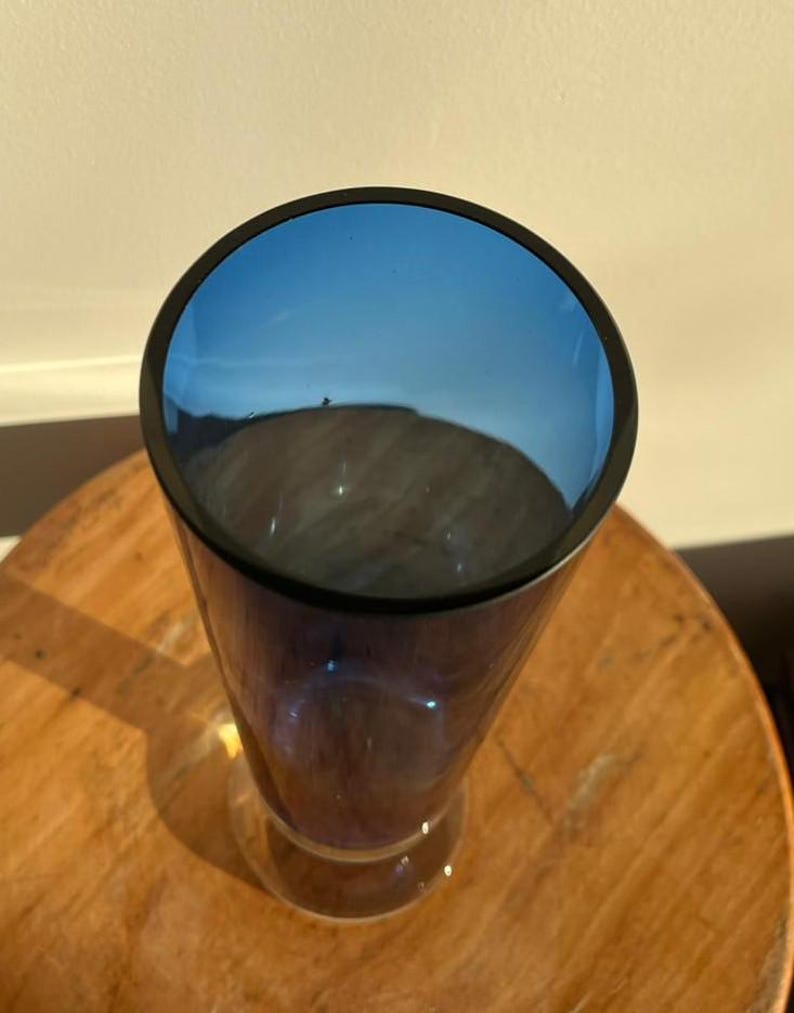 Op de afbeelding: Een hoge, heldere glazen vaas met een uitlopende voet en een donkerblauwe rand. De binnenkant van de vaas is diepblauw. De vaas staat op een houten oppervlak.