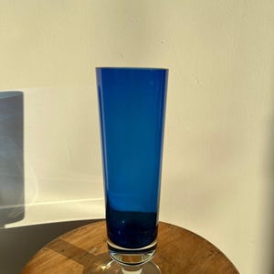 Op de afbeelding: Een hoge, kobaltblauwe glazen vaas met een heldere, cirkelvormige basis. De vaas heeft een slanke, cilindrische vorm en staat op een houten oppervlak. Het glas heeft een diepe, rijke blauwe kleur en de vaas is leeg.