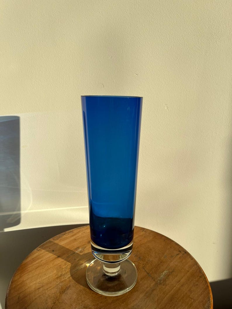 Op de afbeelding: Een hoge, kobaltblauwe glazen vaas met een heldere, cirkelvormige basis. De vaas heeft een slanke, cilindrische vorm en staat op een houten oppervlak. Het glas heeft een diepe, rijke blauwe kleur en de vaas is leeg.