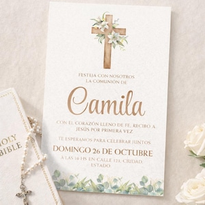 Op de afbeelding: Een crèmekleurige uitnodigingskaart met een houten kruis versierd met witte bloemen. De kaart bevat de naam "Camila" in gouden schrift en evenementdetails in het Spaans. Een witte Bijbel en rozenkrans zijn zichtbaar.