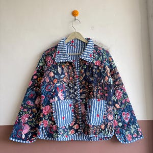 Könnte beinhalten: Eine gesteppte Jacke mit dunkelblauem Grund und einem lebendigen Blumenmuster in Rosa, Rot und Grün. Das Kleidungsstück hat einen Kragen und zwei Vordertaschen mit blau-weißem vertikalem Streifenmuster, das mit kleinen roten Herzen verziert ist.