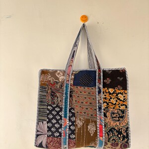May include: A patchwork tote bag with a variety of patterned fabrics in shades of blue, brown, and red. The bag has a striped handle and trim, and is hanging on a wall. The bag is rectangular and appears to be quilted.