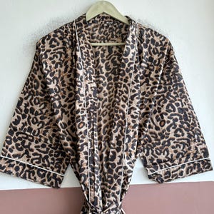 May include: A long-sleeved robe featuring a brown and black leopard print pattern throughout. The garment includes a white piped trim along the front opening, collar, and sleeve cuffs, and comes with a matching fabric tie belt at the waist.