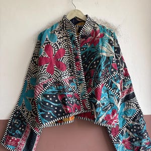 May include: A quilted jacket featuring a patchwork of floral and geometric patterns in vibrant shades of pink, turquoise, black, and white. The garment has long sleeves, a straight hem, and black and white striped trim along the edges.