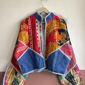 May include: A cropped jacket featuring a patchwork design of various colorful fabrics, including red, blue, and orange floral and paisley patterns. The garment has a high collar, long sleeves, and white stitched detailing along the edges.