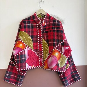 Könnte beinhalten: Eine kurze Steppjacke mit rot-schwarzem Karomuster, kombiniert mit floralen und fruchtigen Drucken. Das Kleidungsstück verfügt über einen Stehkragen, lange Ärmel sowie eine rot-weiß karierte Einfassung an den Kanten und Taschen.