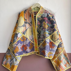 May include: A cropped jacket made from quilted fabric featuring a patchwork of floral and botanical patterns in shades of yellow, purple, orange, and black. The garment has long sleeves, a straight hem, and yellow and white striped trim.