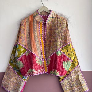 May include: A cropped jacket made from quilted fabric featuring a patchwork of various floral and geometric patterns in shades of orange, pink, yellow, and green. The garment has long sleeves, a stand-up collar, and purple and white checkered trim.