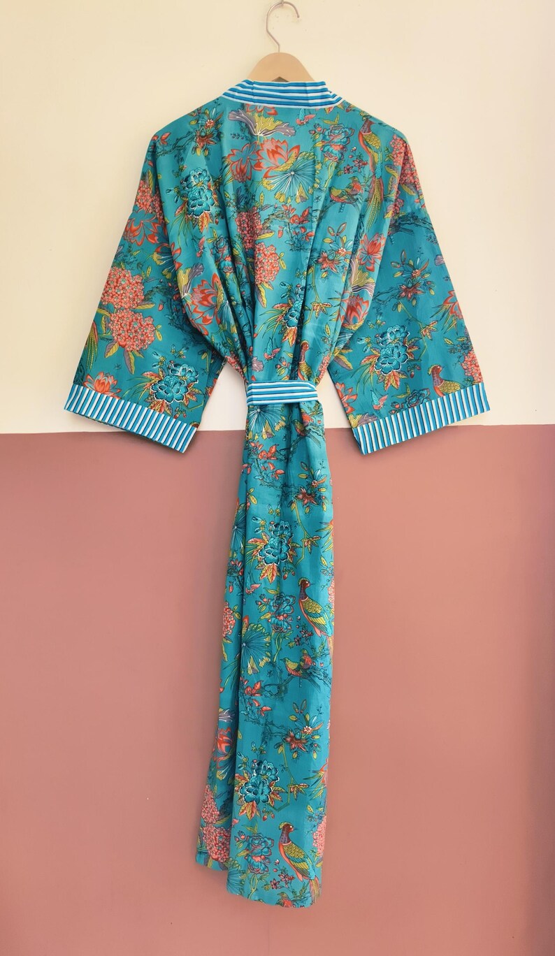 May include: A long-length robe in a vibrant teal blue featuring a colorful floral and bird print. The garment includes wide sleeves with white and blue striped cuffs, a matching tie belt at the waist, and a coordinating striped collar trim.