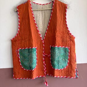 May include: A sleeveless waistcoat featuring a quilted orange fabric with a white and red striped trim along the edges. The garment includes two green patterned patch pockets on the front and a tie closure, with a light-coloured quilted interior lining.