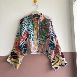 Peut inclure: Une veste courte présentant un patchwork vibrant de motifs floraux et botaniques dans des tons de rouge, vert, bleu et jaune. Le vêtement possède de longues manches larges, un col montant et des coutures visibles sur tout le tissu matelassé.