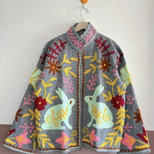 Könnte beinhalten: Graue Jacke mit farbenfrohen Blumen- und Hasenstickereien. Die Jacke hat einen hohen Kragen und ein Rautenmuster. Die Stickerei umfasst rote, gelbe, rosa und grüne Akzente, die ein lebendiges und detailliertes Design schaffen.