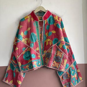 May include: A cropped jacket featuring a vibrant pink base with a repeating pattern of teal floral motifs and yellow leaves. The garment has a mandarin collar, wide sleeves, and white and red striped trim along the edges and seams.