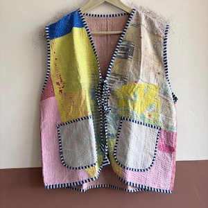 Puede incluir: Un chaleco sin mangas hecho de tela acolchada con un diseño de retazos en paneles amarillos, rosas, azules y blancos. La prenda tiene dos bolsillos delanteros, un cuello en V y un cierre de lazo frontal, todo con un borde a rayas azul y blanco.