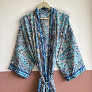 May include: A long-sleeved robe featuring a vibrant paisley pattern in shades of turquoise, blue, and tan. The garment has a shawl collar, wide sleeves, and a matching fabric tie belt at the waist, all constructed from a smooth, lightweight fabric.