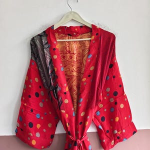 May include: A red robe featuring a multicolored polka dot pattern with blue, yellow, and grey circles. The garment has wide sleeves, a tie waist, and an interior lining displaying a gold-toned fabric with a repeating geometric pattern.