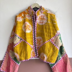 May include: A cropped jacket featuring a mustard yellow base with white and orange circular floral patterns. The garment includes long sleeves with pink and green patchwork sections, a high collar, and a black and white patterned trim along the front opening.