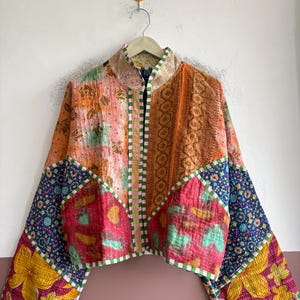May include: A cropped jacket constructed from patchwork fabric featuring a variety of colorful patterns including floral, geometric, and abstract designs in shades of orange, pink, yellow, blue, and green, finished with a green and white checkered trim.