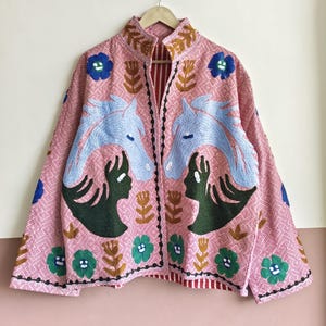 May include: Pink embroidered jacket with a high collar and a geometric pattern. The jacket features light blue horse heads and green human silhouettes facing each other. Additional floral and leaf embroidery in blue, green, and brown.