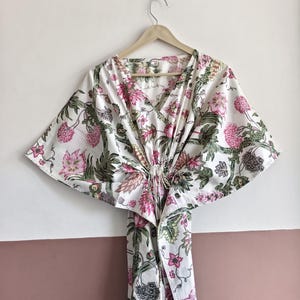 May include: A white floral print kaftan dress with pink and green accents, hanging on a wooden hanger. The dress features a V-neck and a tie at the waist. The floral pattern includes large pink flowers and green leaves.