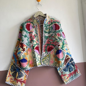 Könnte beinhalten: Eine kurze Jacke mit einem lebendigen Patchwork aus gestickten Blumen- und geometrischen Mustern in Rot-, Blau-, Gelb- und Grüntönen. Das Kleidungsstück hat einen Stehkragen, lange Ärmel und eine helle Basis mit sichtbaren Nähten.