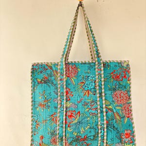 May include: A quilted tote bag with a vibrant floral pattern in shades of turquoise, red, and blue. The bag features long handles and is suspended from a wall. The floral design includes birds and other botanical elements.