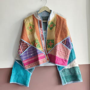 May include: A cropped jacket featuring a patchwork design of various fabrics in shades of orange, yellow, pink, and turquoise. The garment has long, wide sleeves, a mandarin collar, and visible white stitching throughout the quilted fabric panels.