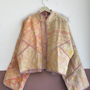 May include: A cropped jacket made from quilted fabric in shades of peach, yellow, and cream with a floral pattern. The garment features a high collar, wide sleeves, and a front opening trimmed with a red and white striped fabric border.