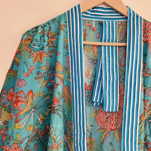 May include: A turquoise kimono robe featuring a vibrant floral and butterfly print in shades of pink, orange, and green. The garment has a contrasting white and blue striped trim along the front opening and collar, hanging on a wooden hanger.