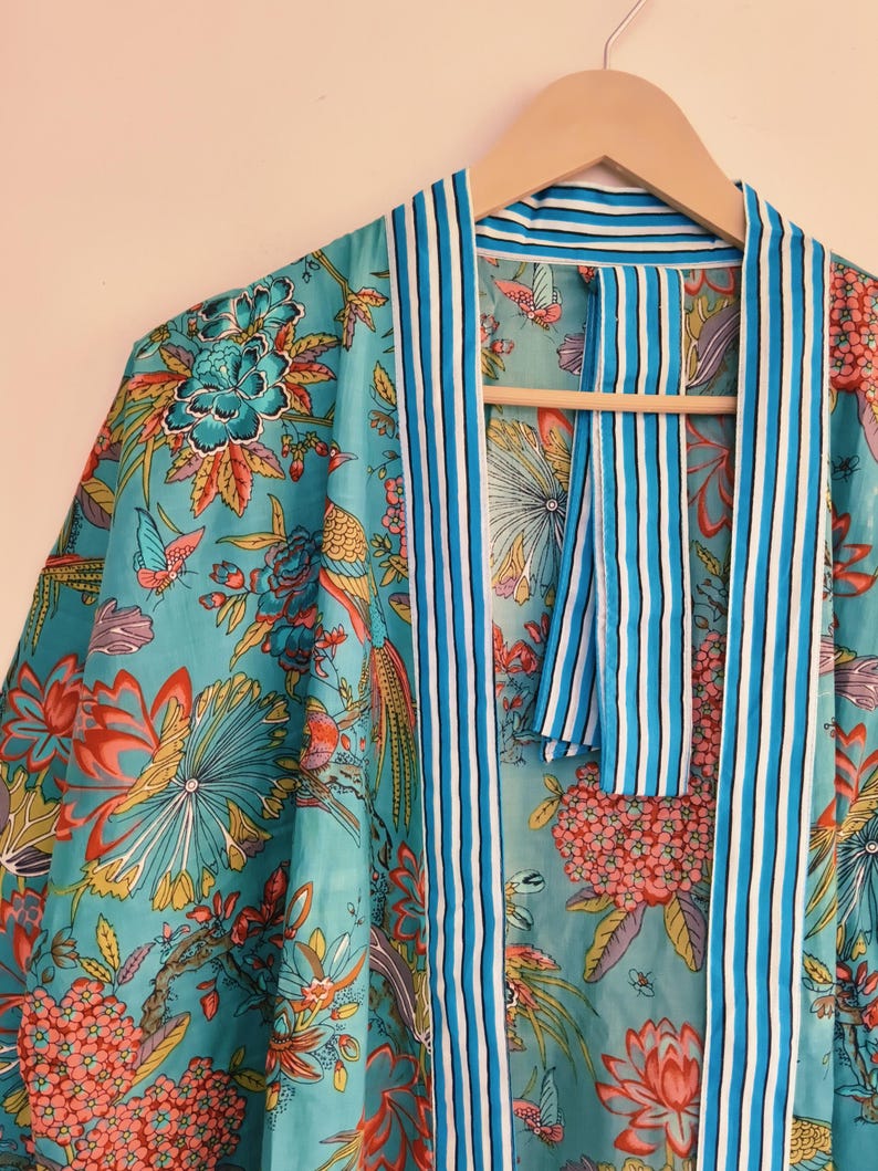 May include: A turquoise kimono robe featuring a vibrant floral and butterfly print in shades of pink, orange, and green. The garment has a contrasting white and blue striped trim along the front opening and collar, hanging on a wooden hanger.