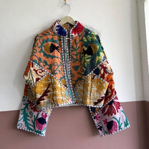 May include: A cropped jacket featuring a vibrant patchwork design with multi-coloured floral and botanical embroidery. The garment has long, wide sleeves, a high collar, and a light-coloured trim along the edges, showcasing intricate stitching.