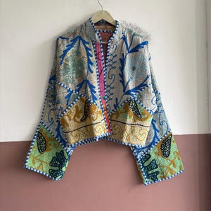 May include: A quilted jacket featuring a patchwork design with various floral and paisley patterns in shades of blue, green, yellow, and white. The garment has long sleeves, an open front, and a mandarin-style collar, all finished with blue trim.