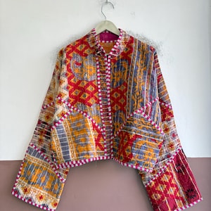 May include: A quilted jacket featuring a vibrant patchwork design in shades of red, yellow, and blue. The garment has long sleeves, a button-front closure, and a collar, with white and pink striped trim along the edges and seams.