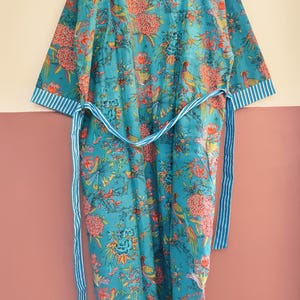 May include: A turquoise robe featuring a vibrant floral and bird pattern in shades of orange, yellow, and green. The garment includes a matching tie belt and white and turquoise striped trim along the collar and sleeve cuffs, hanging on a hanger.