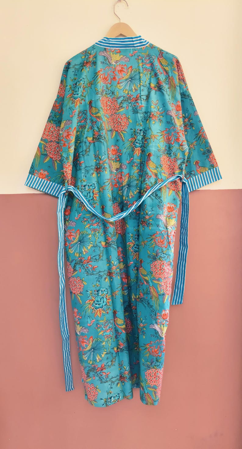 May include: A turquoise robe featuring a vibrant floral and bird pattern in shades of orange, yellow, and green. The garment includes a matching tie belt and white and turquoise striped trim along the collar and sleeve cuffs, hanging on a hanger.
