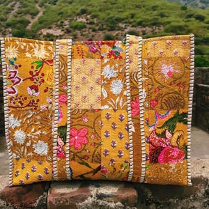 May include: A rectangular tote bag featuring a quilted patchwork design in shades of mustard yellow, gold, and orange. The surface displays various floral and botanical block-print patterns in pink, white, and green, with two striped fabric handles.
