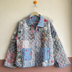 Puede incluir: Una chaqueta acolchada con un diseño de patchwork de patrones florales y de cachemira en gris, azul y blanco. La prenda incluye un cuello y dos bolsillos delanteros de tela a rayas azules y blancas con pequeños corazones rojos.