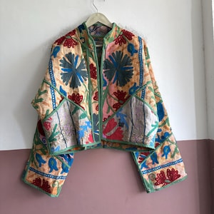 May include: A cropped jacket featuring a patchwork design with intricate floral embroidery in shades of red, blue, and orange. The garment has long sleeves, a stand-up collar, and green fabric trim along the edges, showcasing a quilted texture.