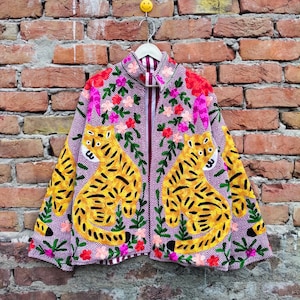 May include: A long-sleeved jacket featuring a purple and white herringbone pattern, adorned with embroidered yellow tigers and colorful floral motifs. The garment has a high collar and an open front design, showcasing detailed stitching throughout.