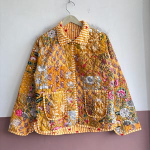 May include: A quilted jacket featuring a vibrant mustard yellow base with a multi-colored floral print. The garment includes a collared neckline, long sleeves, two front patch pockets, and white and yellow striped trim along the edges.