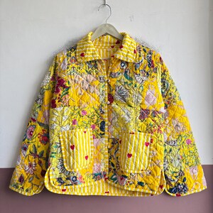 May include: A quilted jacket in a vibrant yellow hue featuring a mix of floral patterns and white striped accents. The garment includes a collared neckline, long sleeves, and two front patch pockets adorned with small red heart embroidery details.