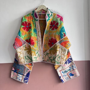 May include: A cropped jacket featuring a vibrant patchwork of multi-coloured floral and geometric embroidery. The garment has long sleeves, an open front, and is finished with a white and brown striped trim along the edges and seams.