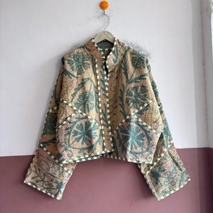 May include: A cropped jacket featuring a quilted texture with a tan base and teal floral patterns. The garment has wide sleeves, a high collar, and white and green checkered trim along the edges, front opening, and sleeve cuffs.
