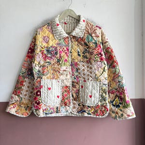 May include: A quilted jacket featuring a patchwork design with various floral and botanical patterns in shades of yellow, pink, and beige. The jacket has a collared neckline, long sleeves, and two front pockets decorated with small red heart embroidery.