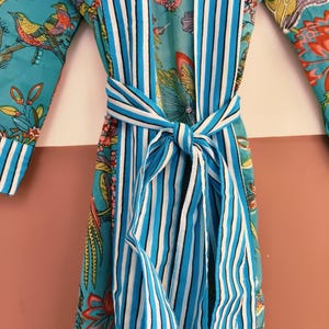 May include: A turquoise robe featuring a vibrant floral and bird pattern in shades of pink, yellow, and green. The garment includes a wide front placket and a matching tie belt, both decorated with vertical white, navy blue, and light blue stripes.