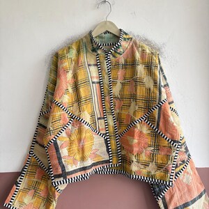 May include: A cropped jacket made from quilted fabric featuring a yellow and orange plaid pattern with floral motifs. The garment has a high collar, wide sleeves, and is finished with black and white striped trim along the edges and seams.