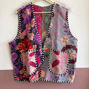 Könnte beinhalten: Eine ärmellose Weste mit Patchwork-Design und lebendiger Blumenstickerei. Die linke Seite ist schwarz mit violetten und roten Blumen, während die rechte Seite hellblau mit bunten Mustern ist. Sie verfügt über zwei Vordertaschen und einen schwarz-weiß gestreiften Saum.