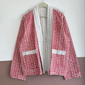 May include: A long-sleeved jacket featuring a red and white diamond pattern with visible running stitch details. The garment has a wide white collar, an open front, and two rectangular front pockets with a white and red geometric print.