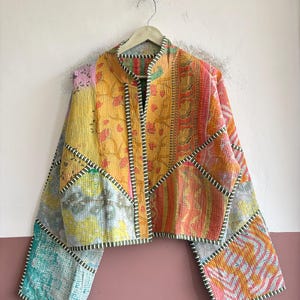 May include: A cropped jacket made from quilted patchwork fabric featuring a variety of floral and geometric patterns in shades of yellow, orange, pink, and teal. The garment has long sleeves, a stand collar, and dark green striped trim.